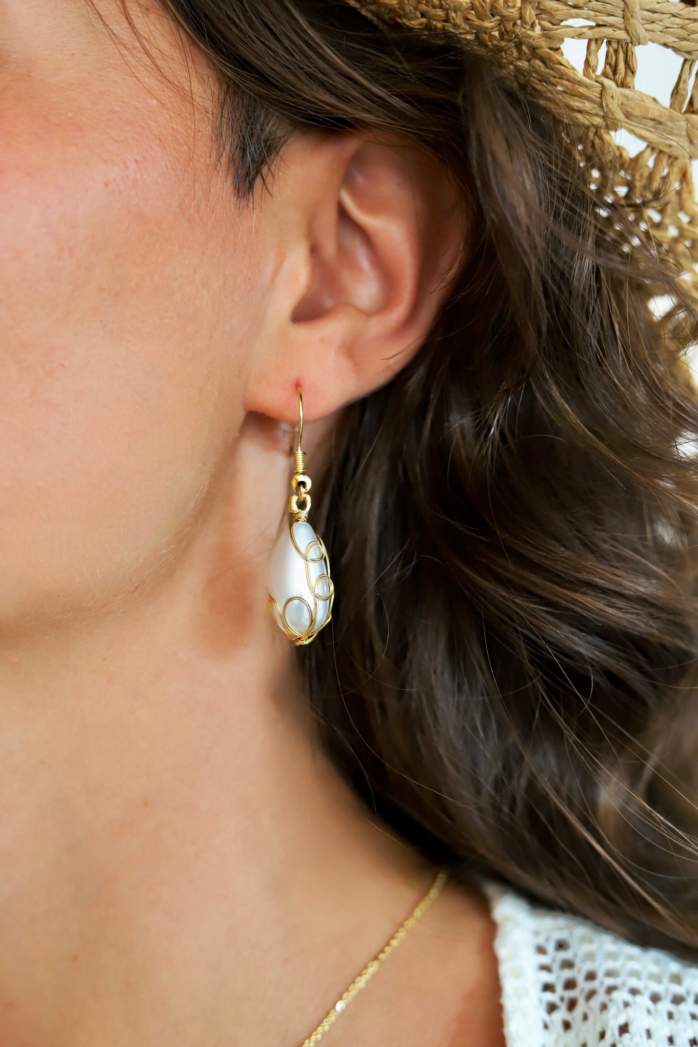 A close-up of a woman's ear wearing a gold drop earring with a white teardrop-shaped shell inlay, next to her brown hair and straw hat.