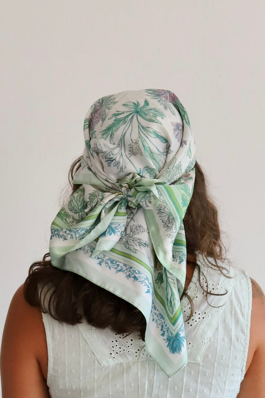 A woman viewed from behind wearing a white scarf with a green and blue botanical pattern tied around her head. She is wearing a white sleeveless top against a plain off-white background.