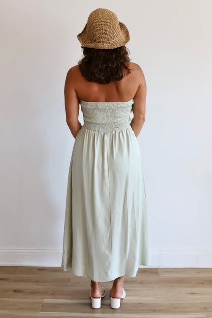 A woman viewed from behind wearing a strapless light sage green midi dress with a smocked bodice, a straw hat, and white heels, standing against a plain white wall on a light wood floor.
