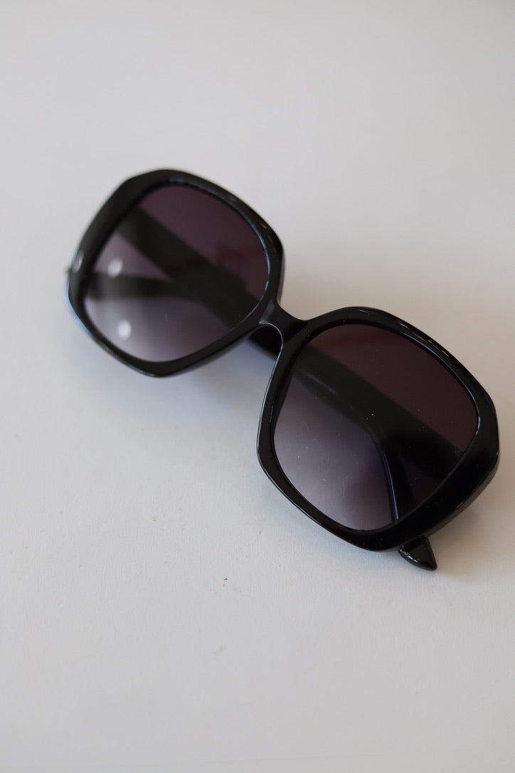 Black oversized square sunglasses with dark gradient lenses, photographed on a plain off-white background.