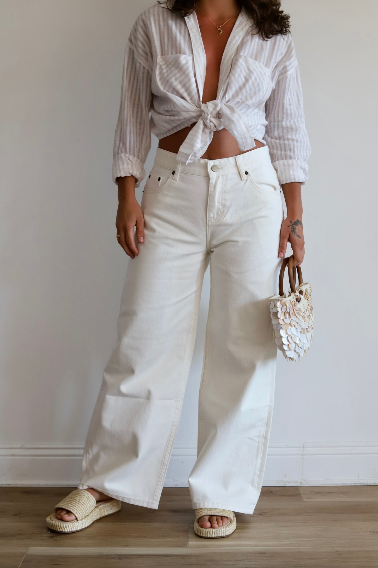 A model wearing a white and light gray vertically striped button-down shirt tied at the waist, paired with high-waisted white wide-leg jeans, beige slide sandals, and holding a small white floral handbag.