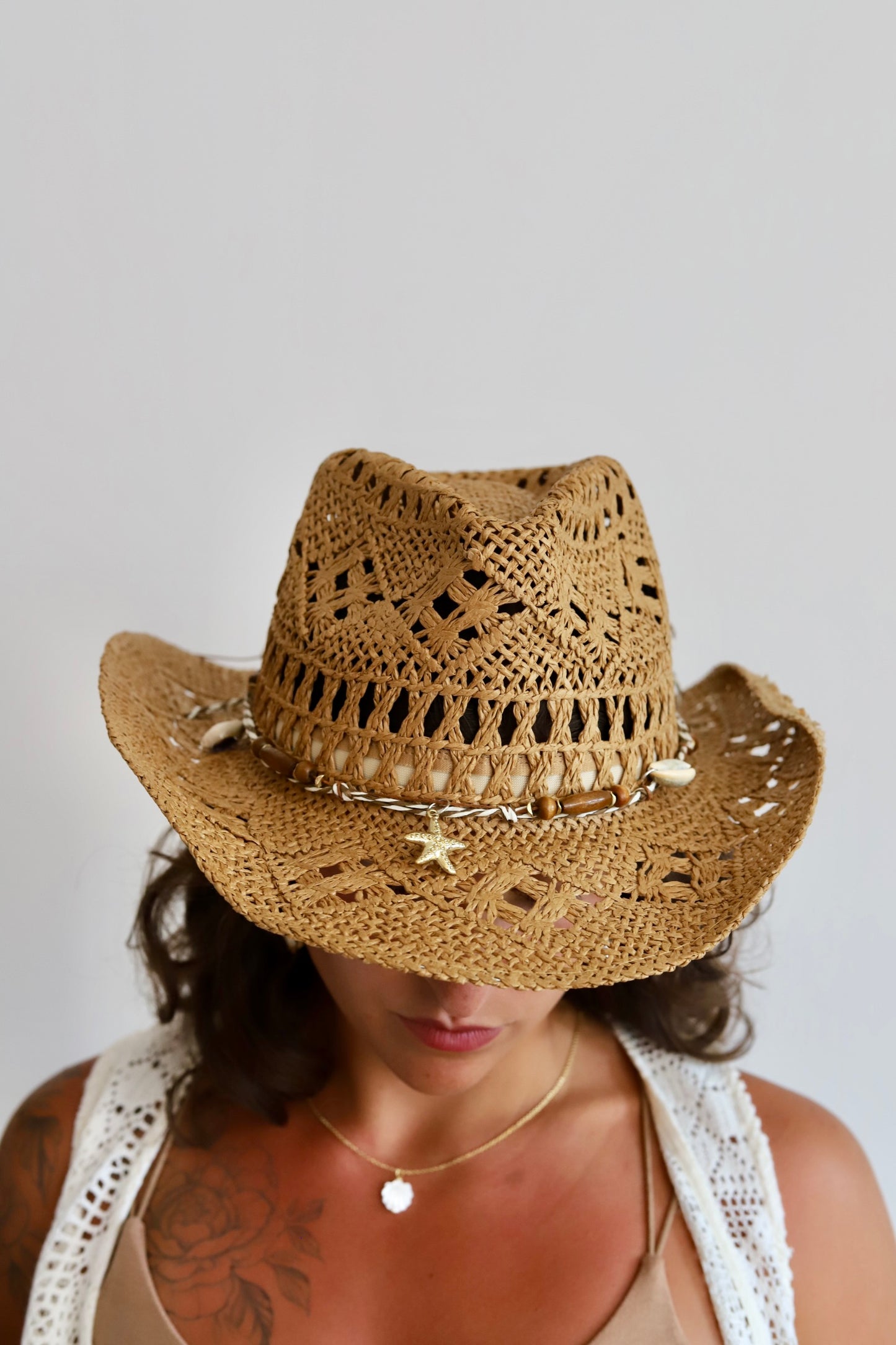 A woman wearing a natural straw cowboy hat with a decorative band of cowrie shells and starfish embellishments, photographed against a plain light gray background.