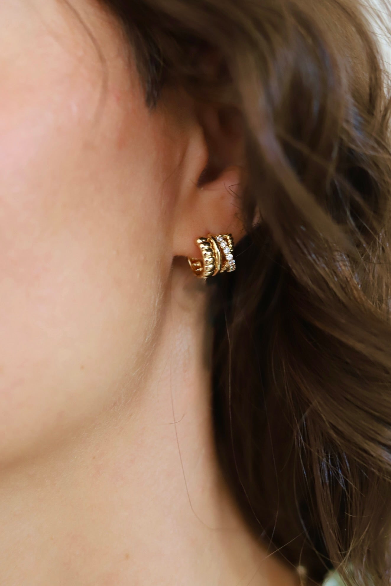 A close-up of a person's ear wearing a small gold huggie hoop earring with textured bands and clear stones.