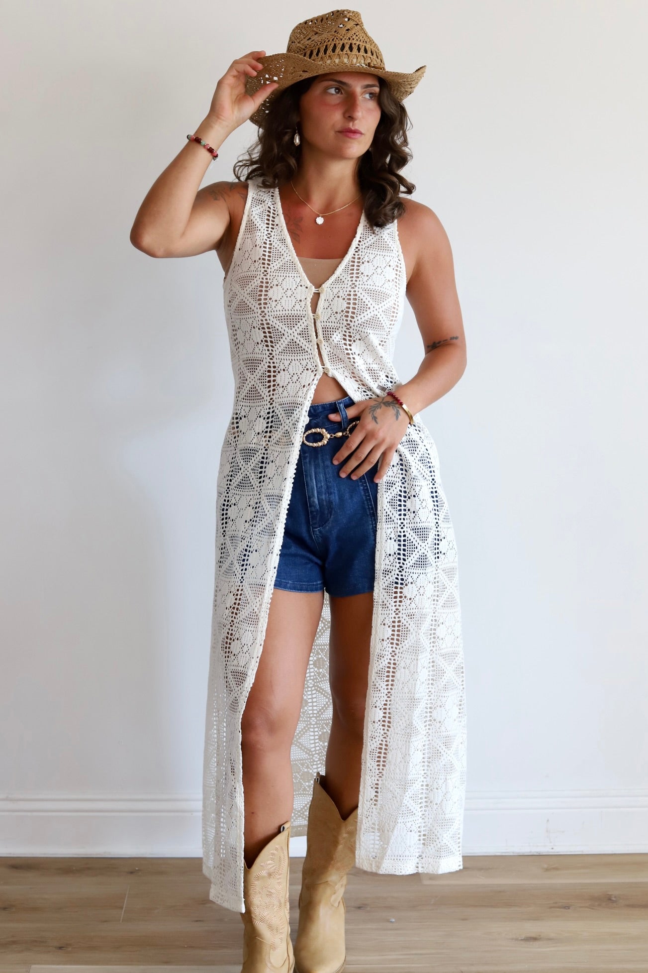 A woman modeling a long white crochet open-front cardigan over a white crop top, paired with denim shorts, a straw cowboy hat, and tan cowboy boots, standing against a plain white wall.