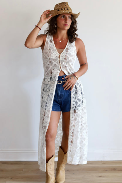 A woman modeling a long white crochet open-front cardigan over a white crop top, paired with denim shorts, a straw cowboy hat, and tan cowboy boots, standing against a plain white wall.