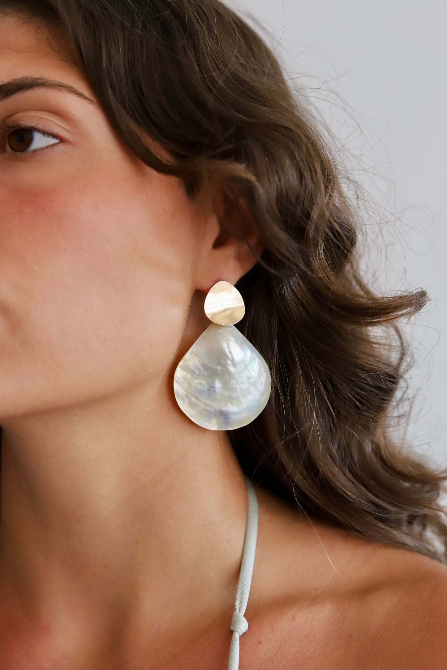 A close-up of a woman wearing a large teardrop-shaped dangle earring made of iridescent mother-of-pearl and a small round gold-toned stud.
