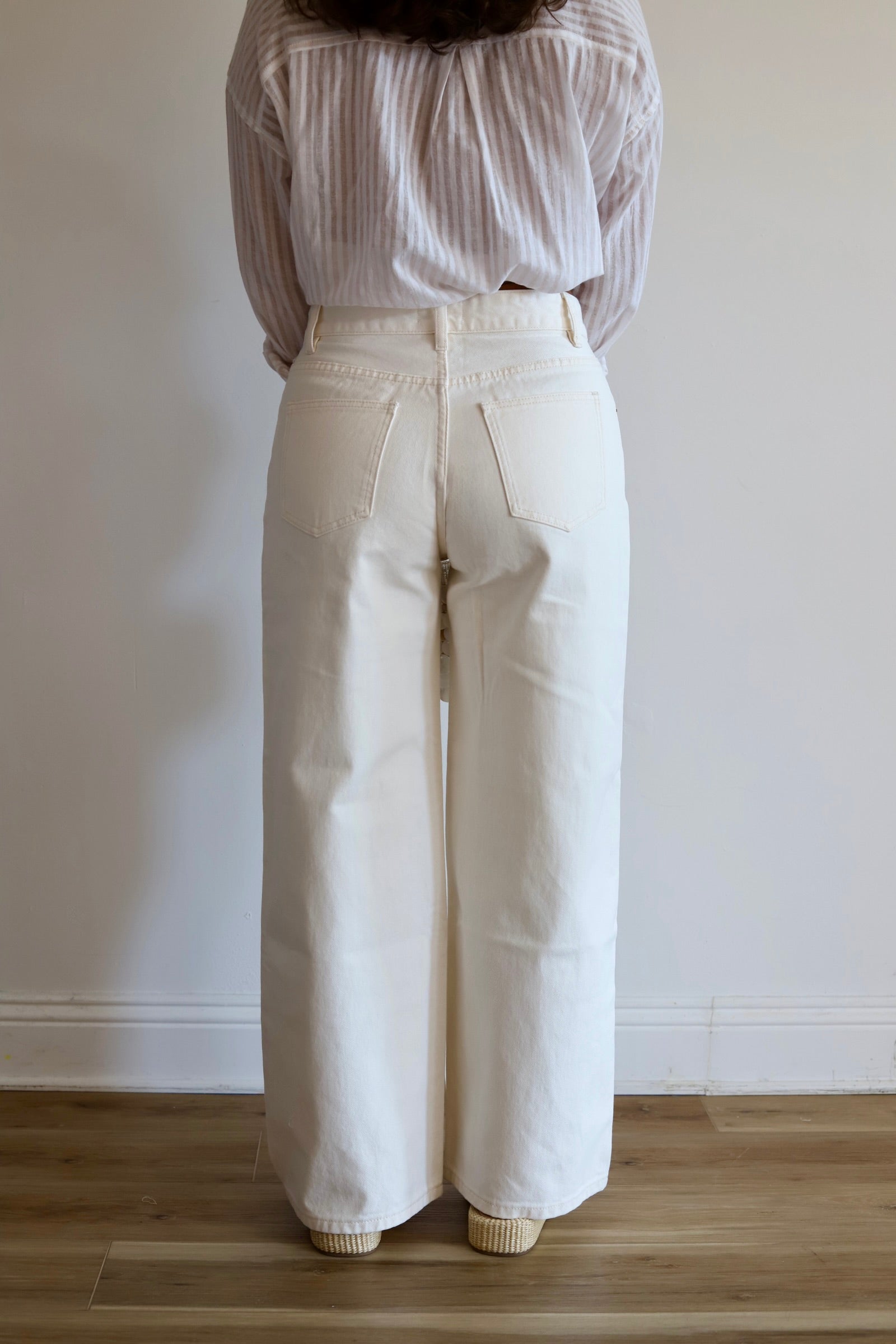 A person shown from the back wearing off-white wide-leg pants and a light striped blouse, standing on a wooden floor against a plain white wall.
