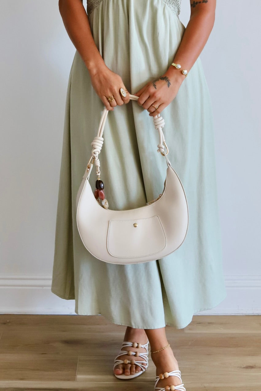 A person wearing a long sage green dress holds a cream-colored crescent-shaped leather shoulder bag with a knotted rope handle. The person is also wearing white strappy flat sandals, standing against a white wall on a light wood floor.