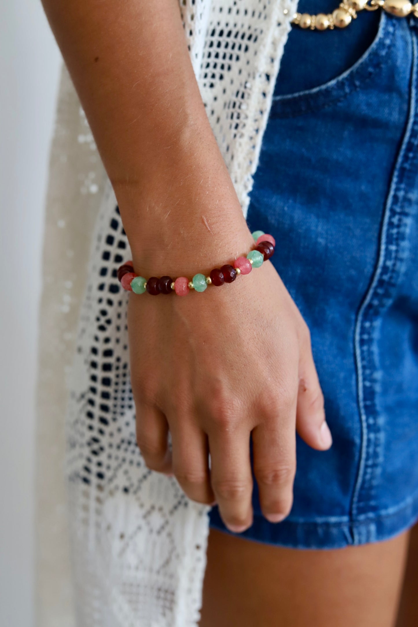 A close-up of a person's wrist wearing a beaded stretch bracelet with faceted beads in burgundy, mint green, and coral pink, styled with a white crochet cardigan and blue denim shorts.
