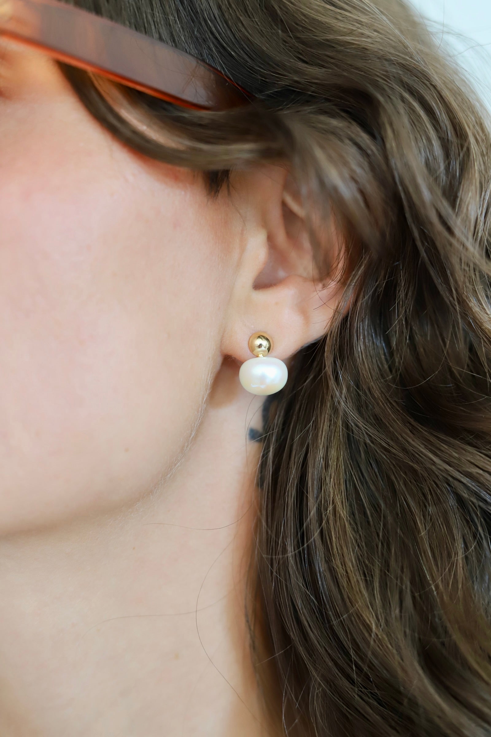 A close-up of a person's ear wearing a small gold stud earring with a white pearl drop, with brown hair and part of a face visible.