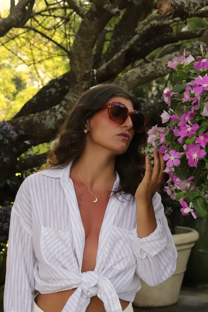 A woman wearing a white and light blue vertically striped button-down shirt tied at the waist, styled with brown sunglasses and a gold crescent necklace, posing outdoors next to pink flowers under a tree.