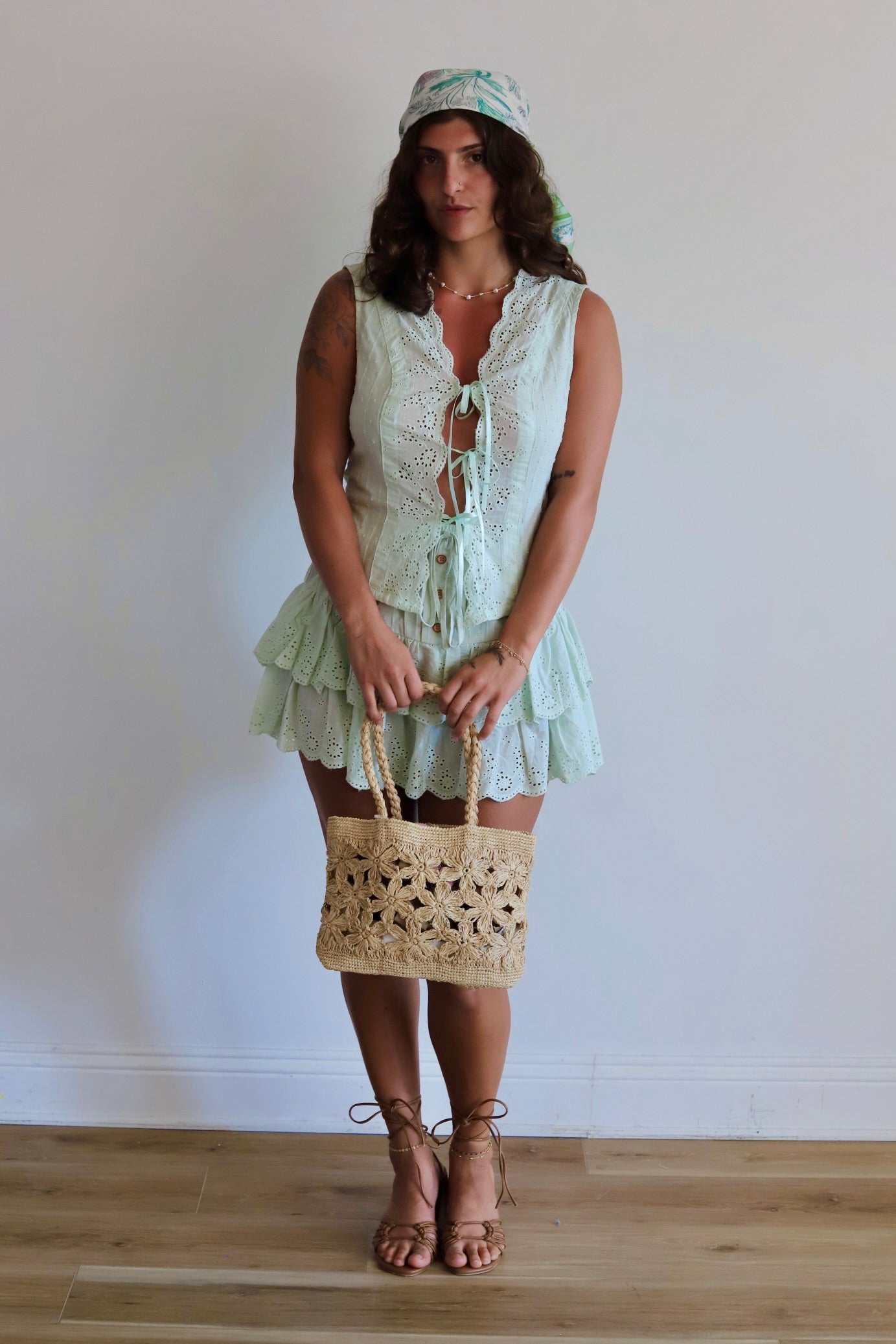 A woman stands in a room with a white wall and light wood floor, wearing a pale mint green lace dress with a matching headscarf, holding a beige woven straw tote bag, and wearing brown strappy high-heeled sandals.