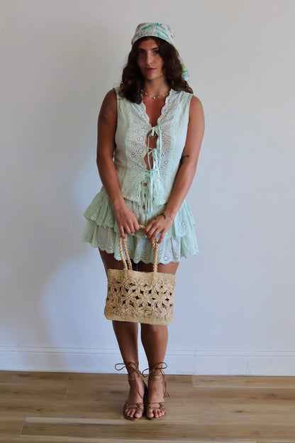 A woman stands in a room with a white wall and light wood floor, wearing a pale mint green lace dress with a matching headscarf, holding a beige woven straw tote bag, and wearing brown strappy high-heeled sandals.