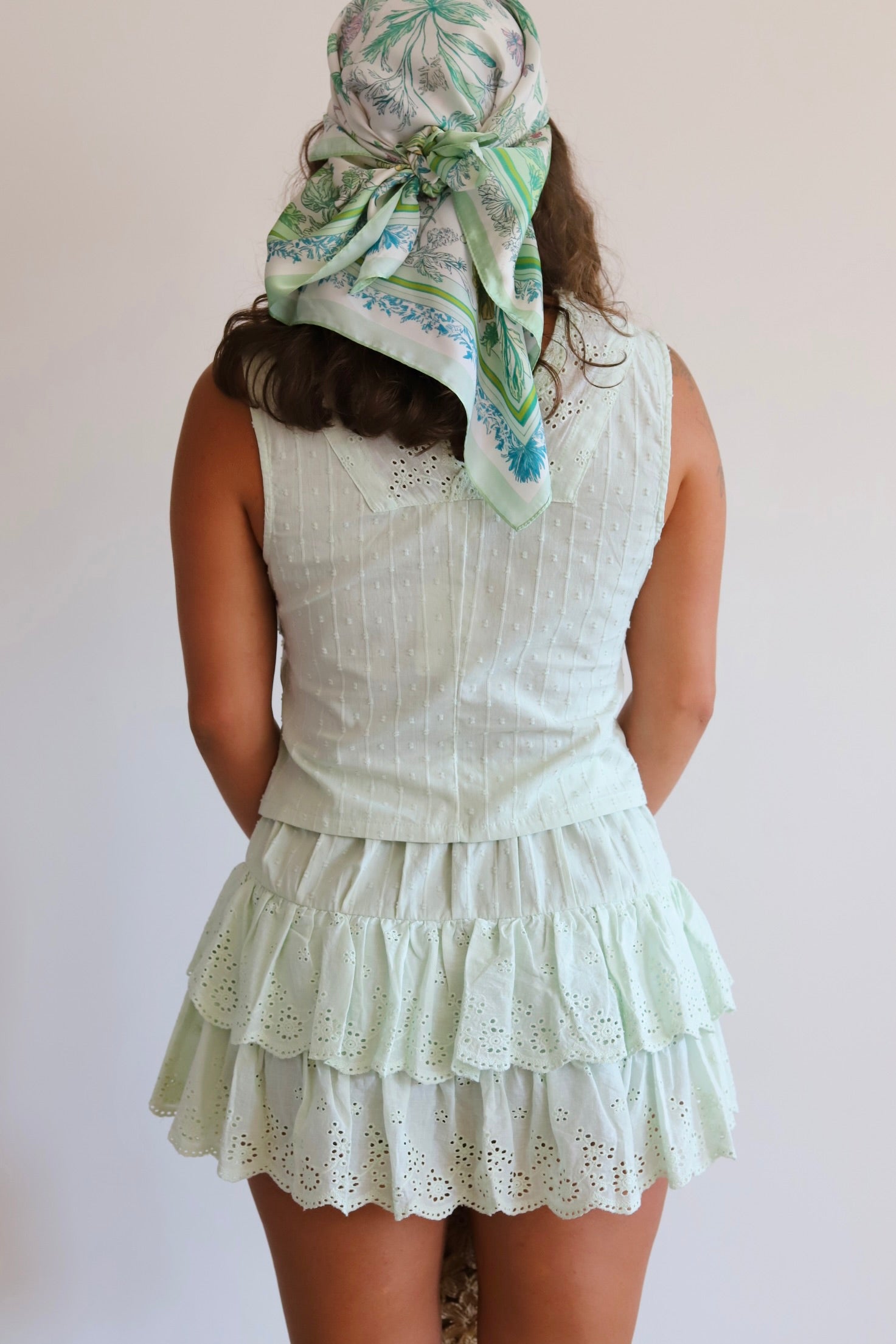 A woman shown from the back wearing a sleeveless light mint green eyelet top and matching tiered ruffle skirt, with a patterned green and white headscarf tied around her hair against a plain light background.