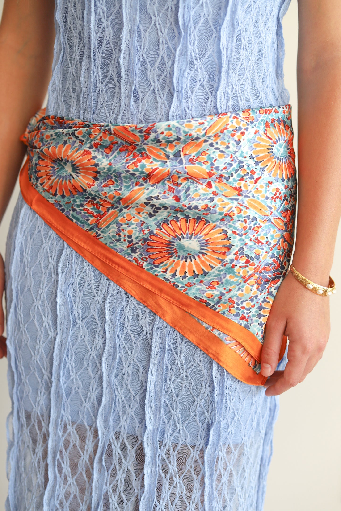 A scarf tied over a lace blue dress. Scarf is shades of blue and orange in a mosaic print style. 