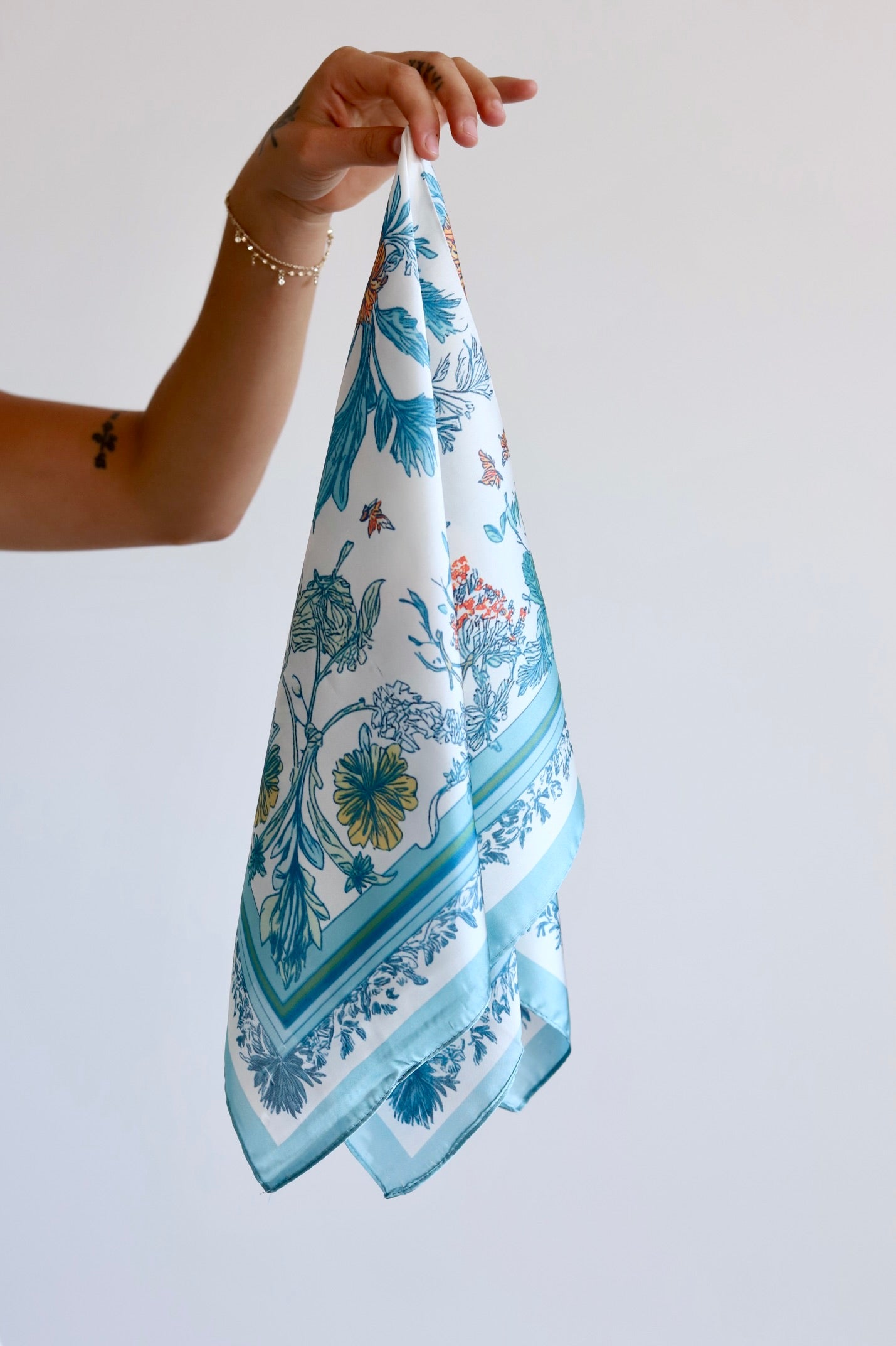 A hand holding a square white scarf with a blue botanical and floral print, featuring a decorative border, against a plain light gray background.