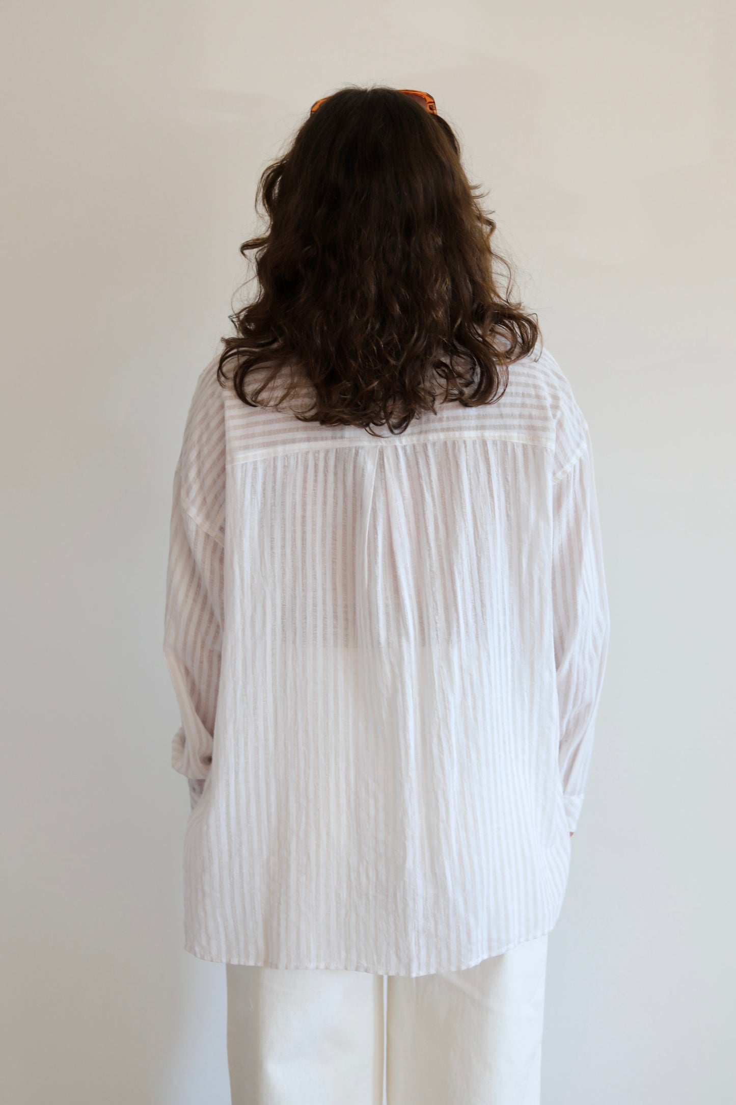 A person viewed from behind wearing a loose white long-sleeve blouse with subtle vertical texture and white pants, standing against a plain light wall.