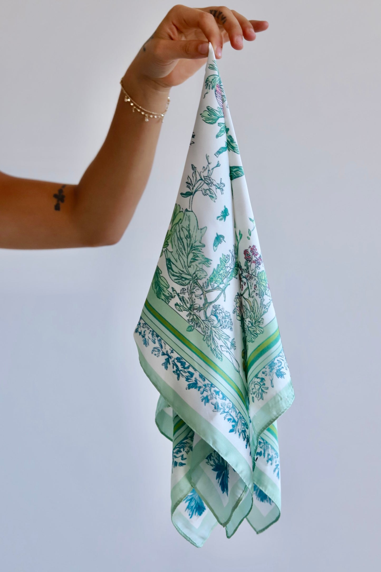 A hand holding a square white scarf with a green botanical and floral toile pattern, set against a plain light gray background.