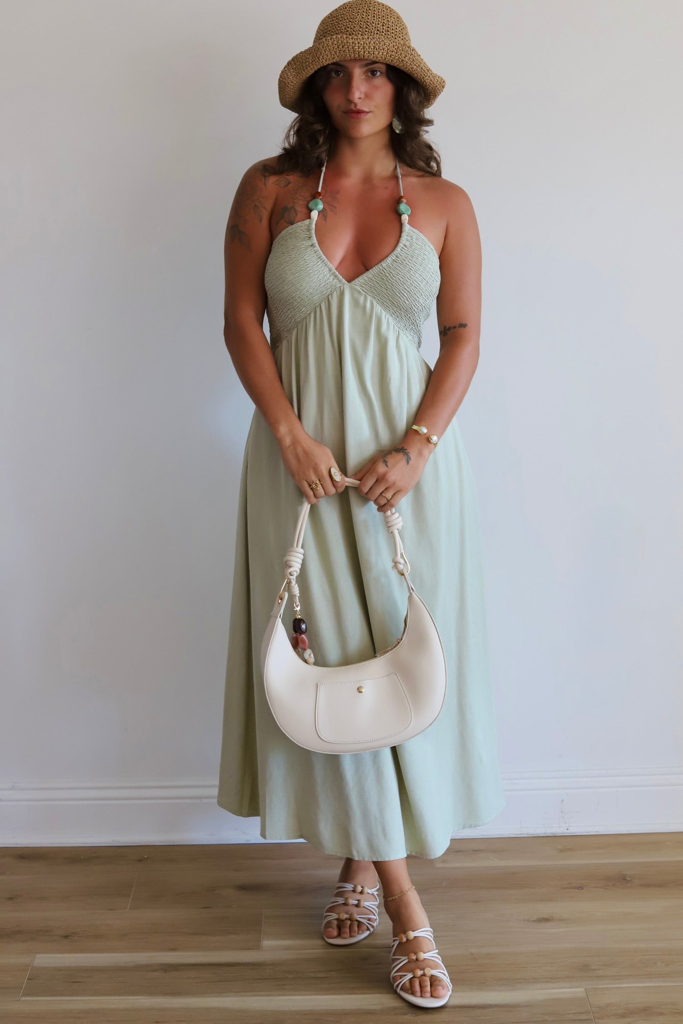 A woman stands indoors wearing a light sage green halter midi dress with a textured bodice and flowing skirt, paired with a straw hat, white sandals, and a white crescent shoulder bag.