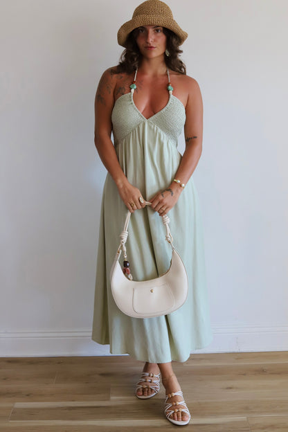 A woman stands indoors wearing a light sage green halter midi dress with a textured bodice and flowing skirt, paired with a straw hat, white sandals, and a white crescent shoulder bag.