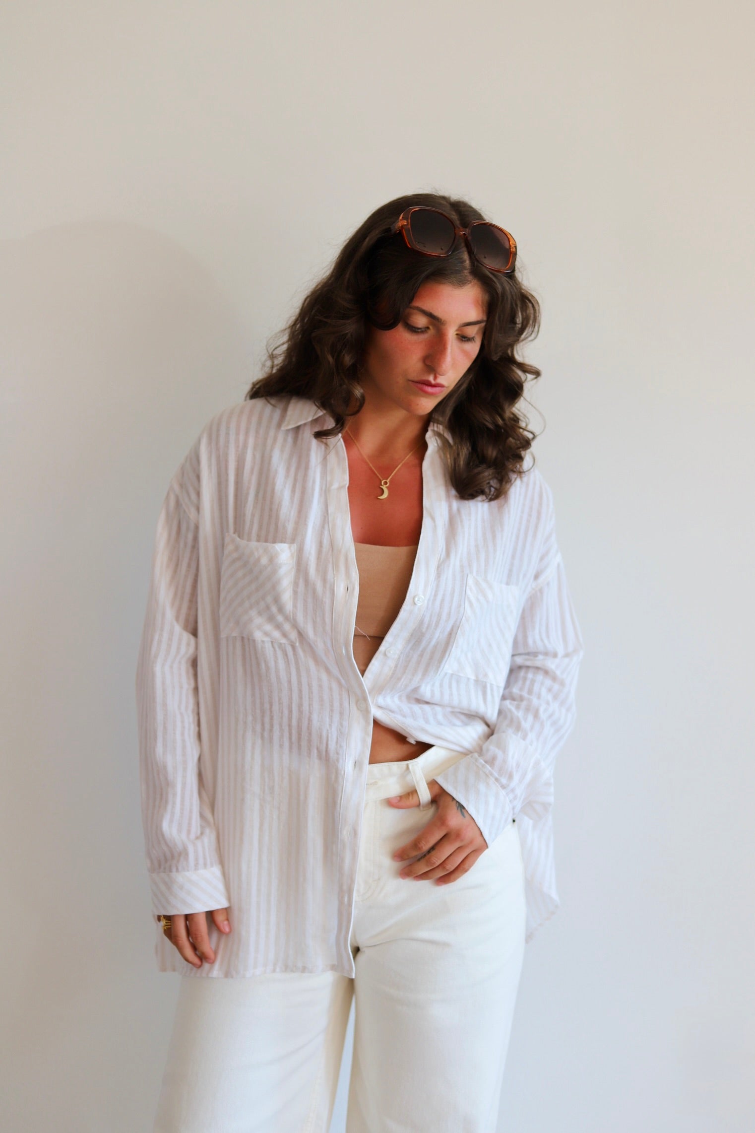 A woman wearing a white long-sleeve button-up shirt with thin vertical pinstripes, styled open over a beige top and paired with white pants, standing against a plain off-white wall.