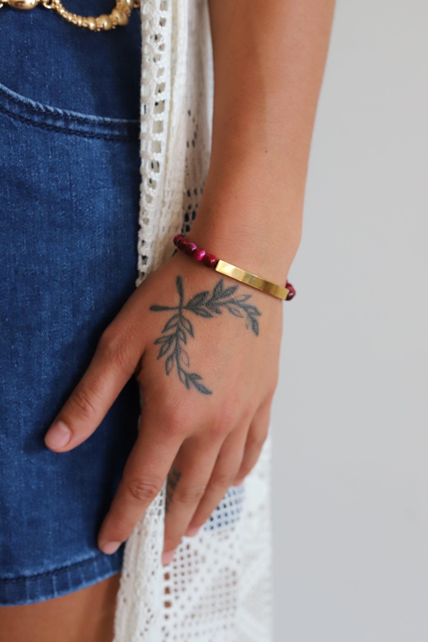 A close-up of a person's hand resting against a white lace garment, wearing a gold bar bracelet and a red beaded bracelet. The person is also wearing blue denim shorts.