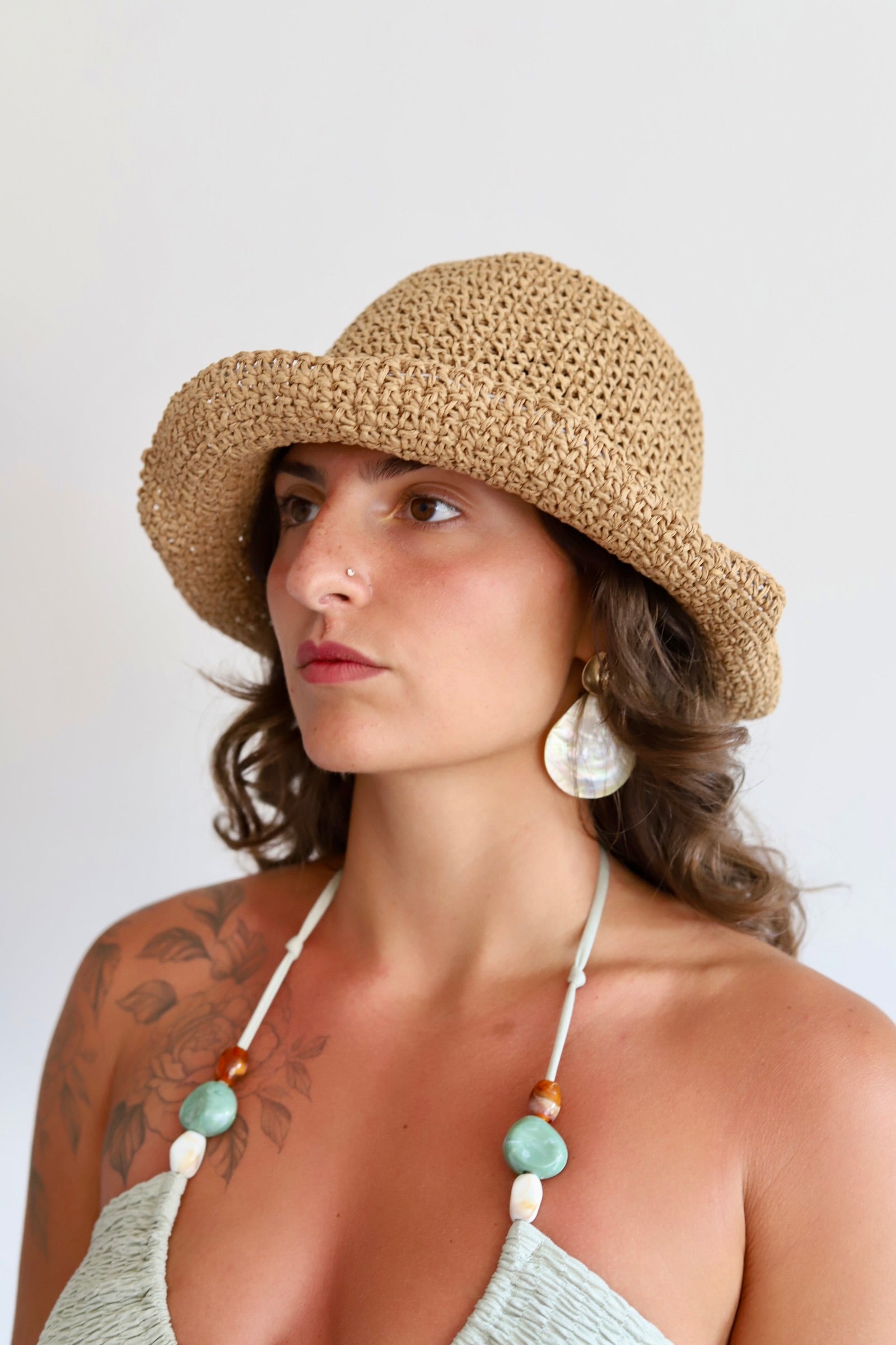 A woman wearing a natural straw sun hat,  and large white shell earrings, shown against a plain light background.