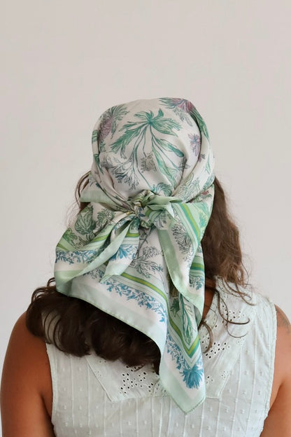 A woman viewed from behind wearing a white scarf with a green and blue botanical pattern tied around her head. She is wearing a white sleeveless top against a plain off-white background.