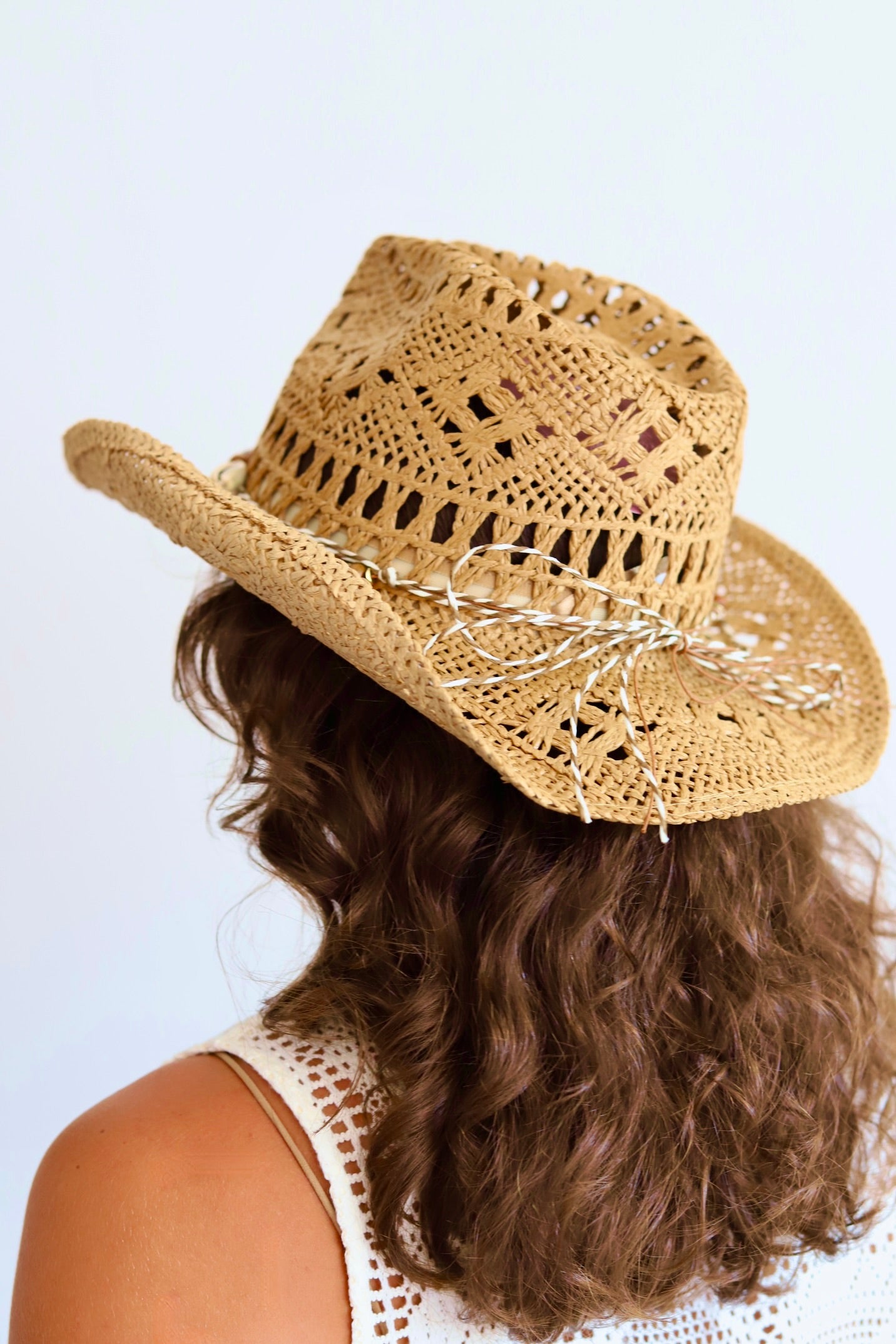 A woman shown from the back wearing a woven straw cowboy hat with a white embroidered bow and brown wavy hair, against a plain light background.