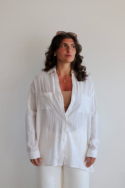 A woman with dark wavy hair wearing a white long-sleeve button-up shirt with thin vertical pinstripes, paired with white pants and sunglasses on her head, standing against a plain light gray wall.